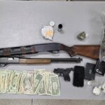 Two rifles, a handgun, stacks of cash, small bags of drugs, and a wallet displayed on a grey table.