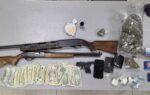 Two rifles, a handgun, stacks of cash, small bags of drugs, and a wallet displayed on a grey table.