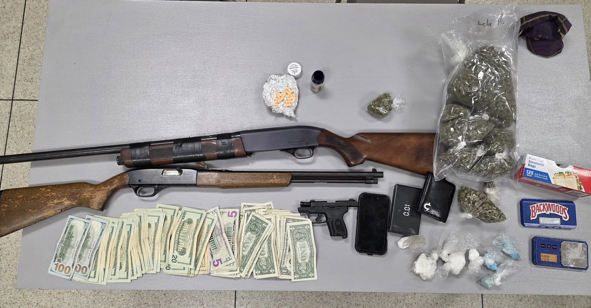 Two rifles, a handgun, stacks of cash, small bags of drugs, and a wallet displayed on a grey table.