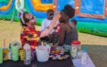 A man in a colorful shirt paints a girl’s face at an outdoor event, with art supplies on the table and inflatable structures in the background.