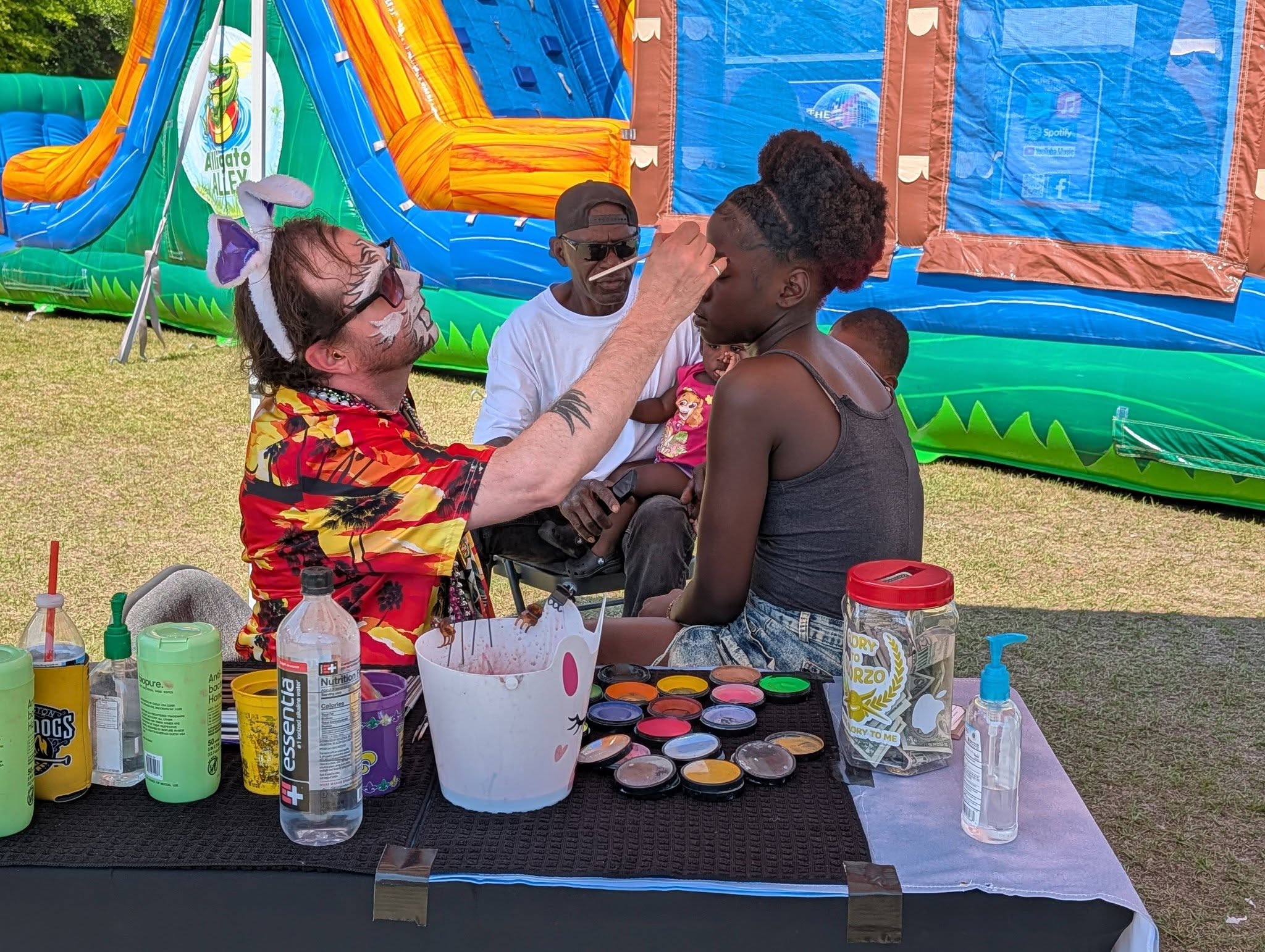 A man in a colorful shirt paints a girl’s face at an outdoor event, with art supplies on the table and inflatable structures in the background.