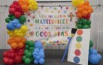 Colorful balloon arch, paint palette prop, and a banner reading "You are God's masterpiece & you are a piece of God's plan" displayed in a decorated indoor setting.