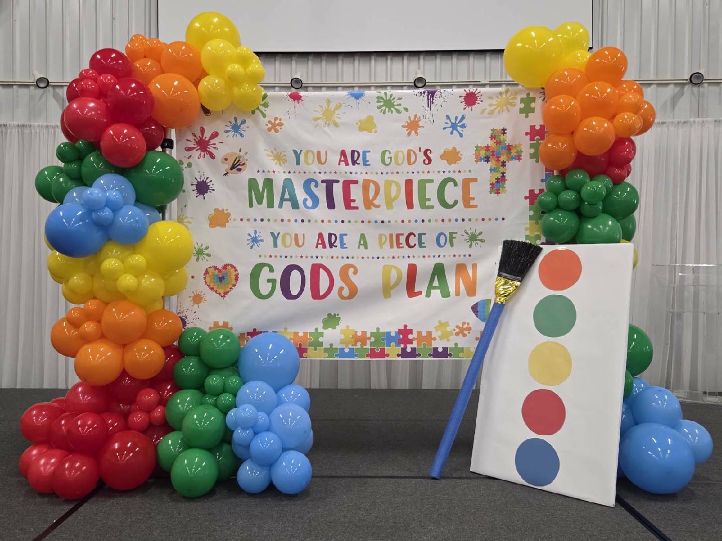 Colorful balloon arch, paint palette prop, and a banner reading "You are God's masterpiece & you are a piece of God's plan" displayed in a decorated indoor setting.