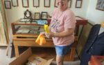 Woman in pink shirt cleaning a display case, holding cleaning supplies, standing next to an open box in a room with framed pictures on the wall.