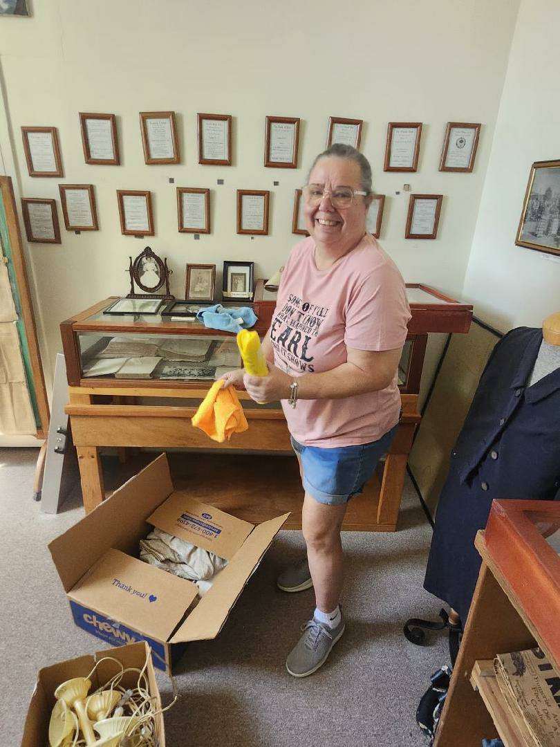 Woman in pink shirt cleaning a display case, holding cleaning supplies, standing next to an open box in a room with framed pictures on the wall.