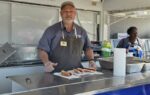 A man wearing an apron and cap stands behind a food counter with three hot dogs placed on the counter in front of him.
