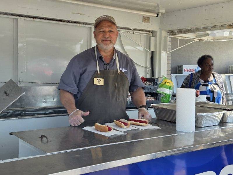 A man wearing an apron and cap stands behind a food counter with three hot dogs placed on the counter in front of him.