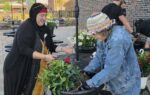 Two women are gardening together outdoors, planting greenery in a large pot. One wears a headscarf, the other a colorful hat and denim jacket.