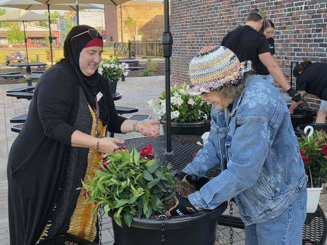 Two women are gardening together outdoors, planting greenery in a large pot. One wears a headscarf, the other a colorful hat and denim jacket.