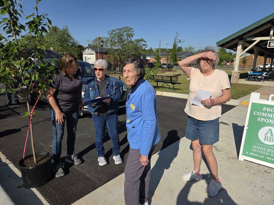 Four older women stand outside on a sunny day, chatting and holding papers, with a tree and building in the background.