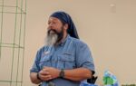 Man with a long beard and blue headscarf stands indoors, wearing a blue shirt and watch, with pet supplies visible in the foreground.