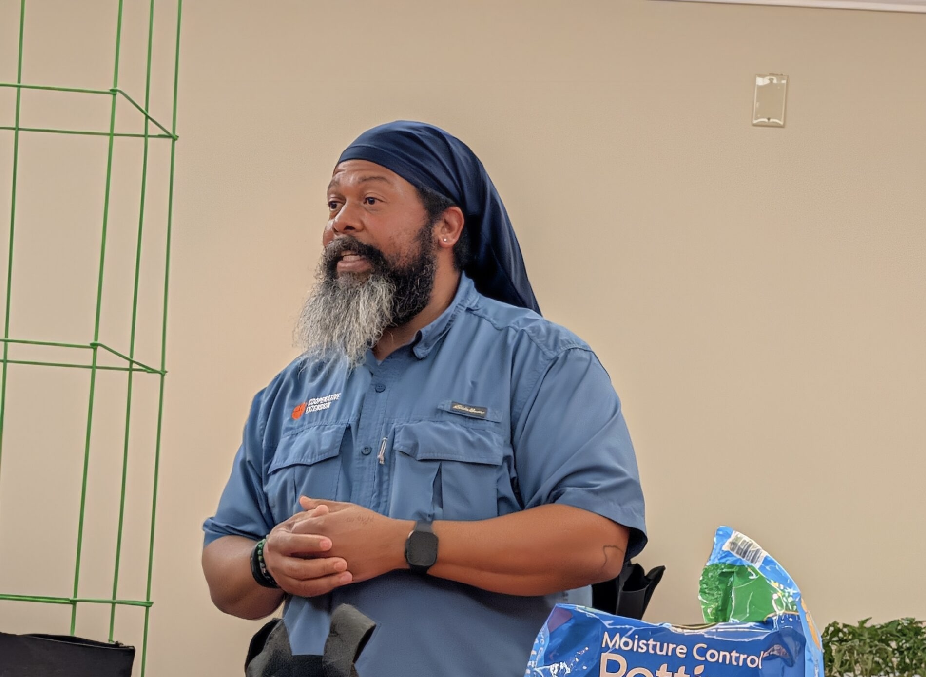 Man with a long beard and blue headscarf stands indoors, wearing a blue shirt and watch, with pet supplies visible in the foreground.