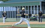 Baseball player in black uniform pitching on a baseball field, building and two people in the background.