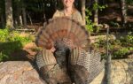 A person in outdoor clothing sits on a log, holding a large wild turkey with fanned tail feathers. A rifle rests against the log nearby.