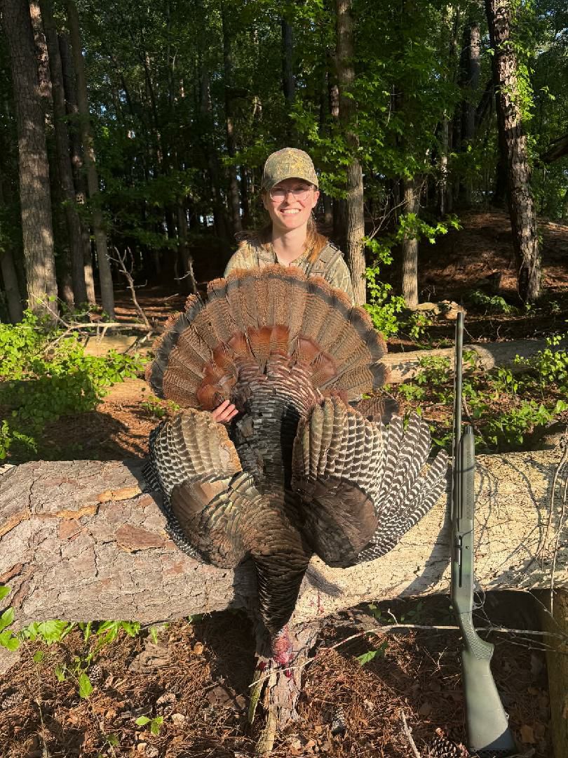 A person in outdoor clothing sits on a log, holding a large wild turkey with fanned tail feathers. A rifle rests against the log nearby.