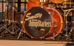Orange drum set with a bass drum labeled "Smitty and the JumpStarters," set up on stage with microphones and other musical equipment visible.