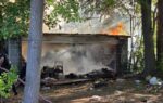 A small building surrounded by trees is on fire, with flames and smoke coming from the roof and debris scattered around the ground.