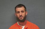 A man with short brown hair and a beard wearing an orange prison jumpsuit stands against a plain gray background, looking at the camera.