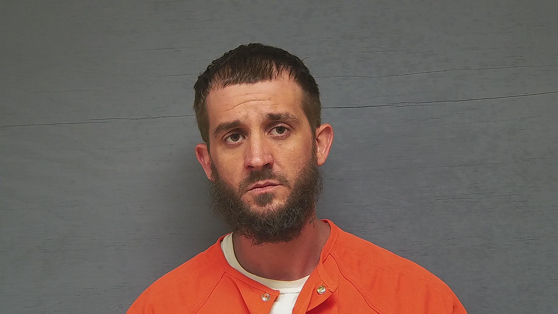 A man with short brown hair and a beard wearing an orange prison jumpsuit stands against a plain gray background, looking at the camera.