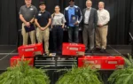 Six people stand on a stage with gift boxes and tool chests in front of them, posing for a photo at an event.