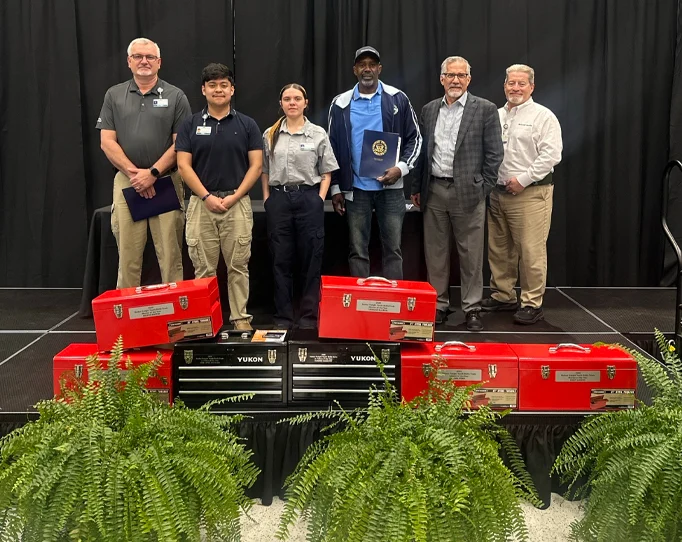 Six people stand on a stage with gift boxes and tool chests in front of them, posing for a photo at an event.