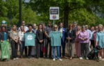 A group of people stands outdoors near a sign that reads "Milele Health." Two people in front hold up green T-shirts. Trees and greenery are in the background.