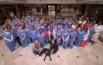 A large group of people, mostly in blue uniforms, pose together indoors. Two dogs and a few people in casual clothing are at the front of the group.