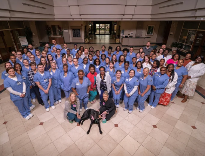 A large group of people, mostly in blue uniforms, pose together indoors. Two dogs and a few people in casual clothing are at the front of the group.