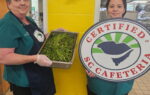 Two cafeteria workers stand indoors, one holding a tray of greens and the other holding a large "Certified SC Cafeteria" sign.