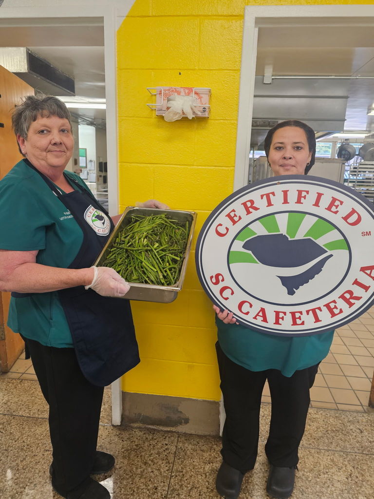 Two cafeteria workers stand indoors, one holding a tray of greens and the other holding a large "Certified SC Cafeteria" sign.