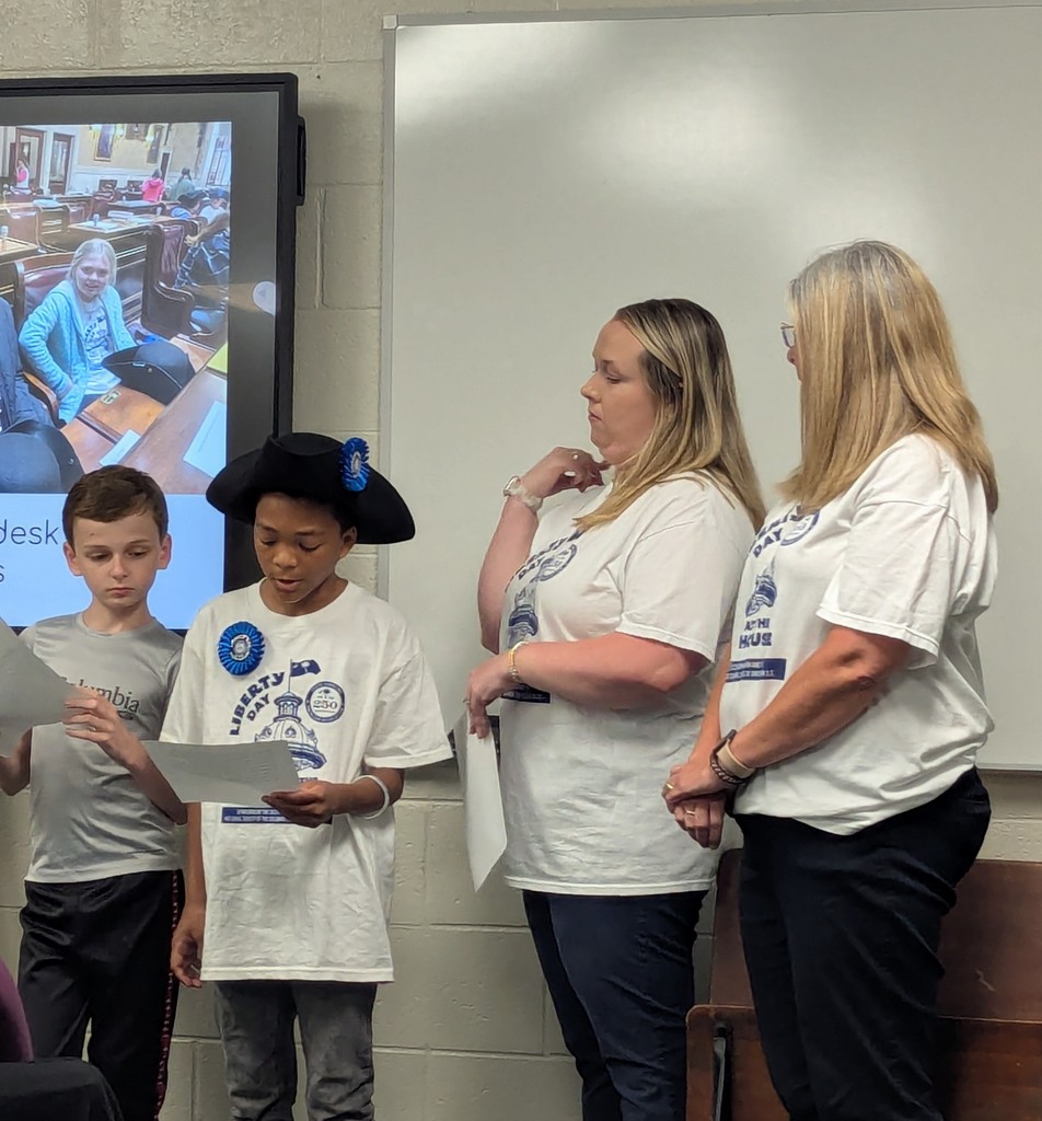 Two boys and two women stand together indoors, with one boy reading from a paper. A projected image appears on the screen behind them.