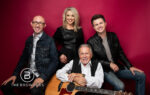 Four smiling people pose in front of a red background, one holding an acoustic guitar, all dressed casually.