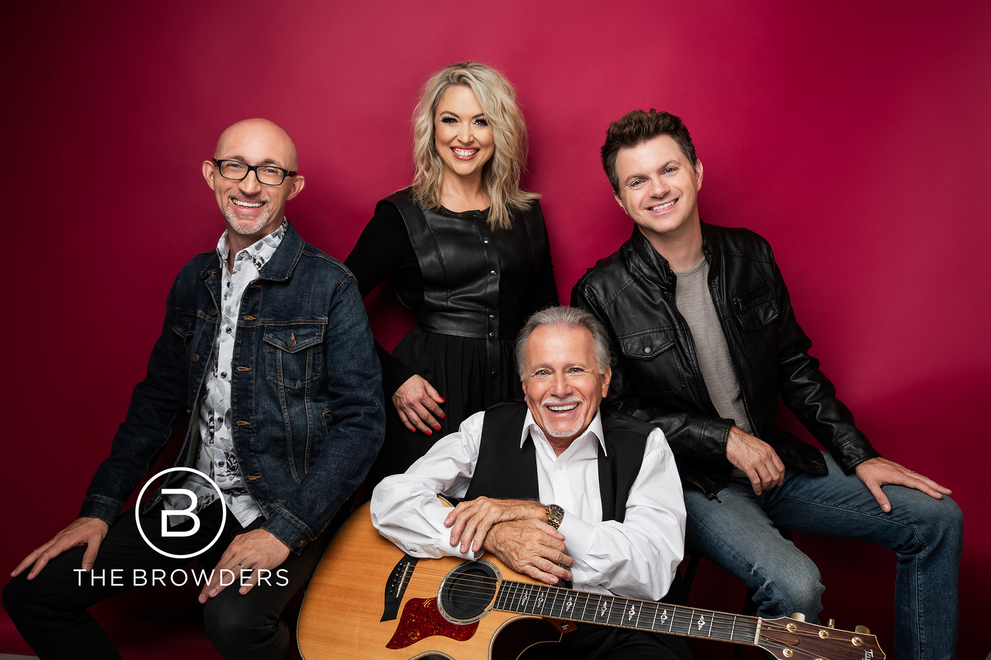 Four smiling people pose in front of a red background, one holding an acoustic guitar, all dressed casually.