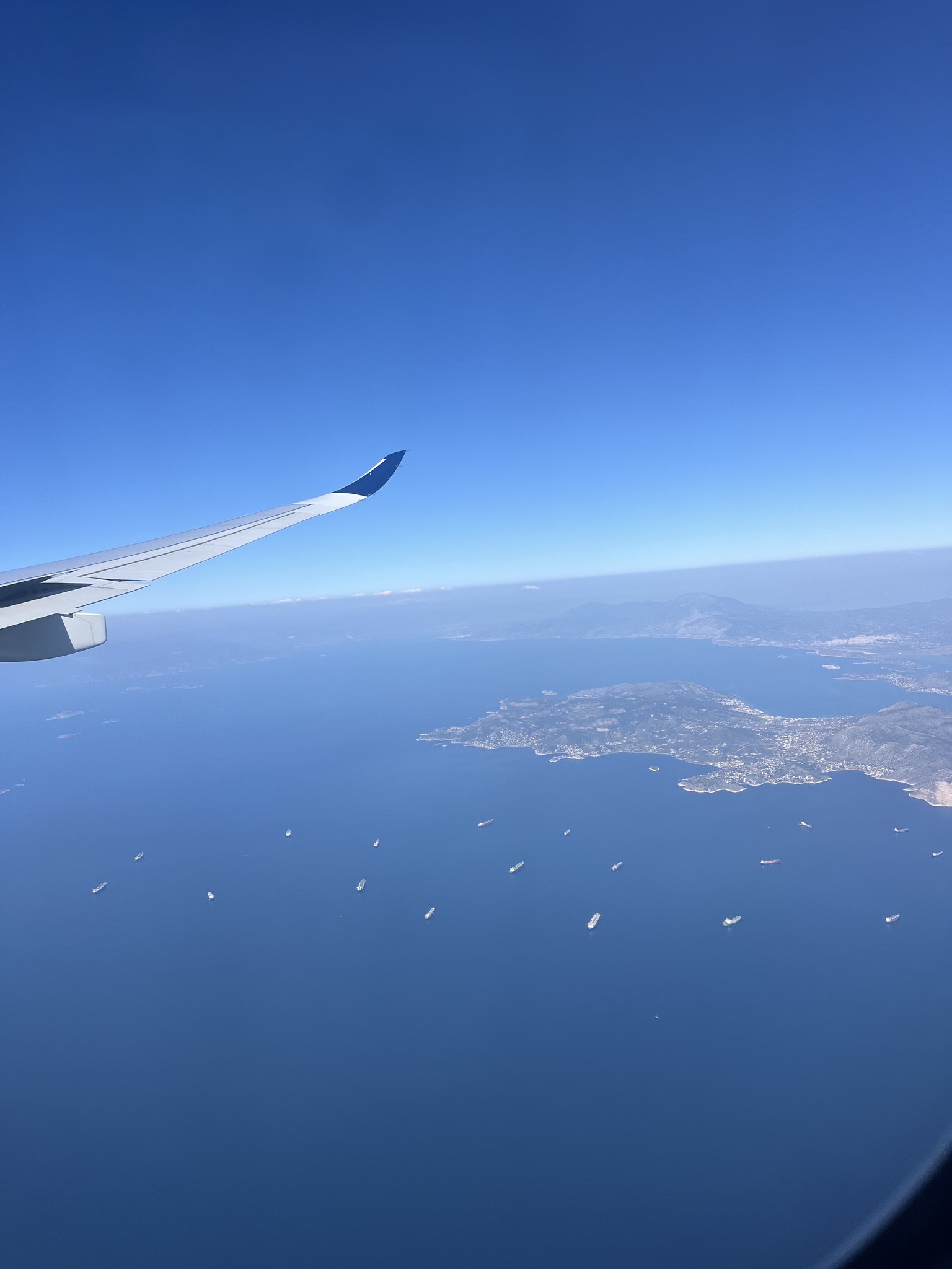 View from airplane window showing wing, blue sky, ocean, scattered boats, and a coastline below.