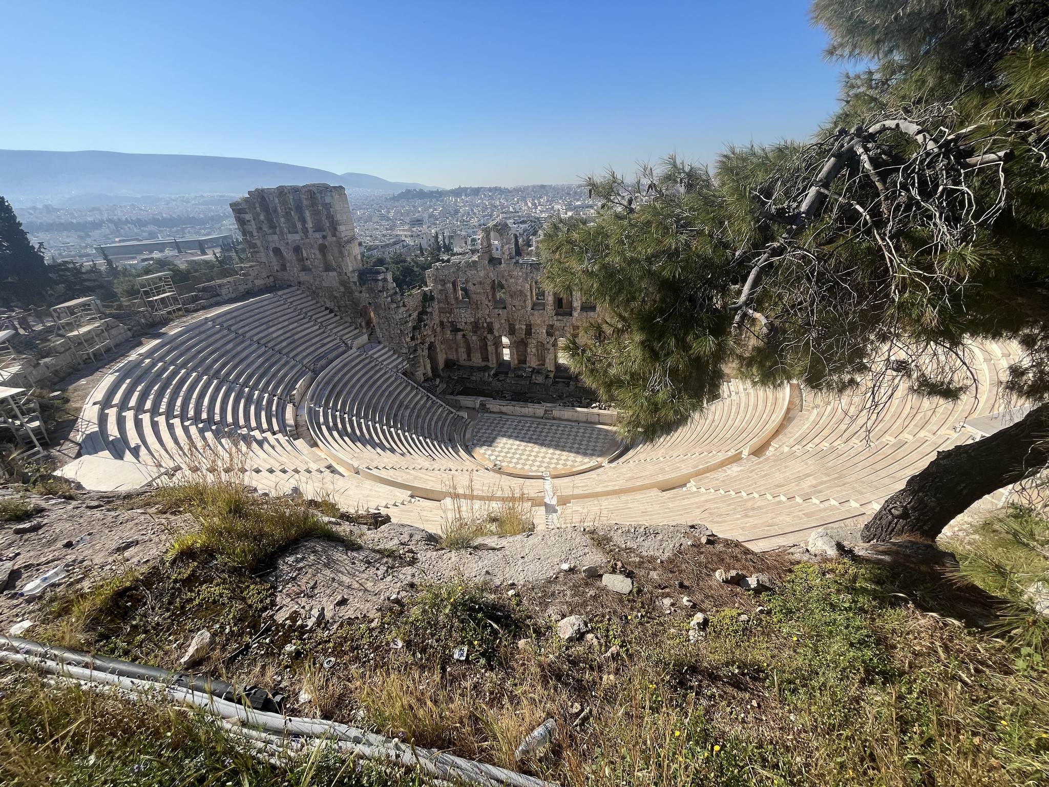 Ancient stone amphitheater with curved seating, partially surrounded by trees, overlooking a city under a clear blue sky.