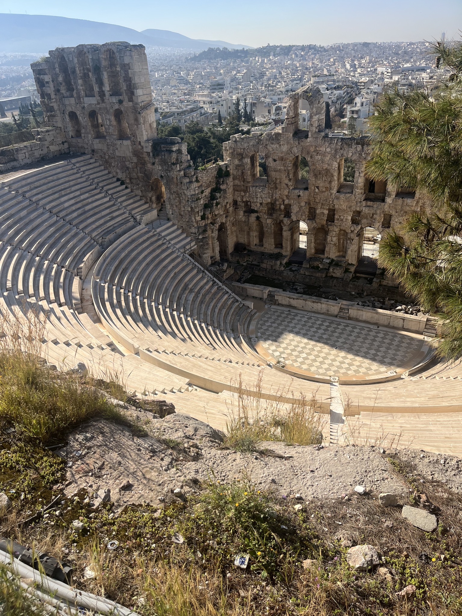Ancient stone amphitheater with semicircular seating, partially ruined stage, surrounded by historical ruins and distant city buildings in the background on a sunny day.