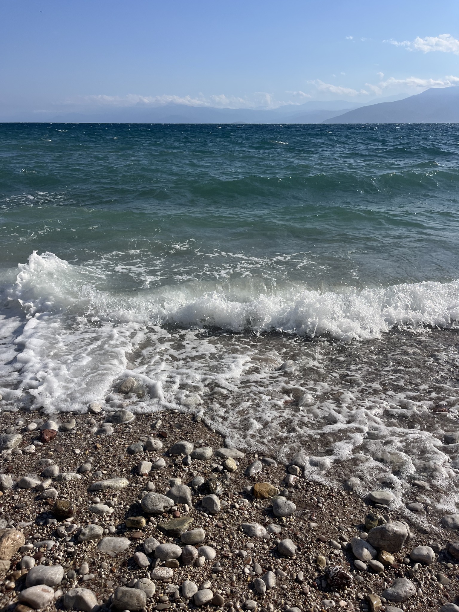 Waves crashing onto a rocky shore under a bright sky, with mountains visible in the distance across the sea.