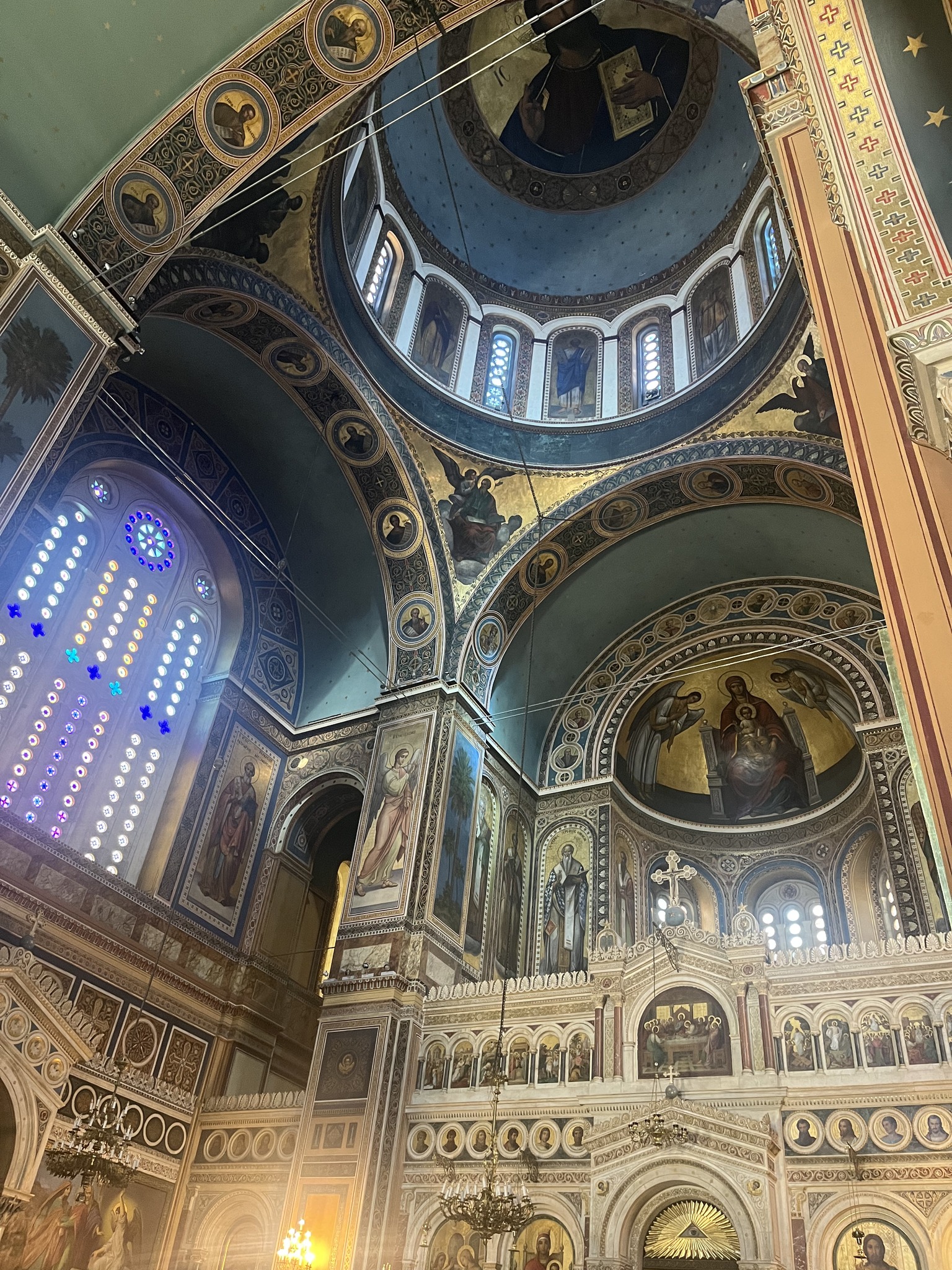 Interior of an ornate church with arched ceilings, detailed frescoes, large stained glass window, and religious artwork above the altar.