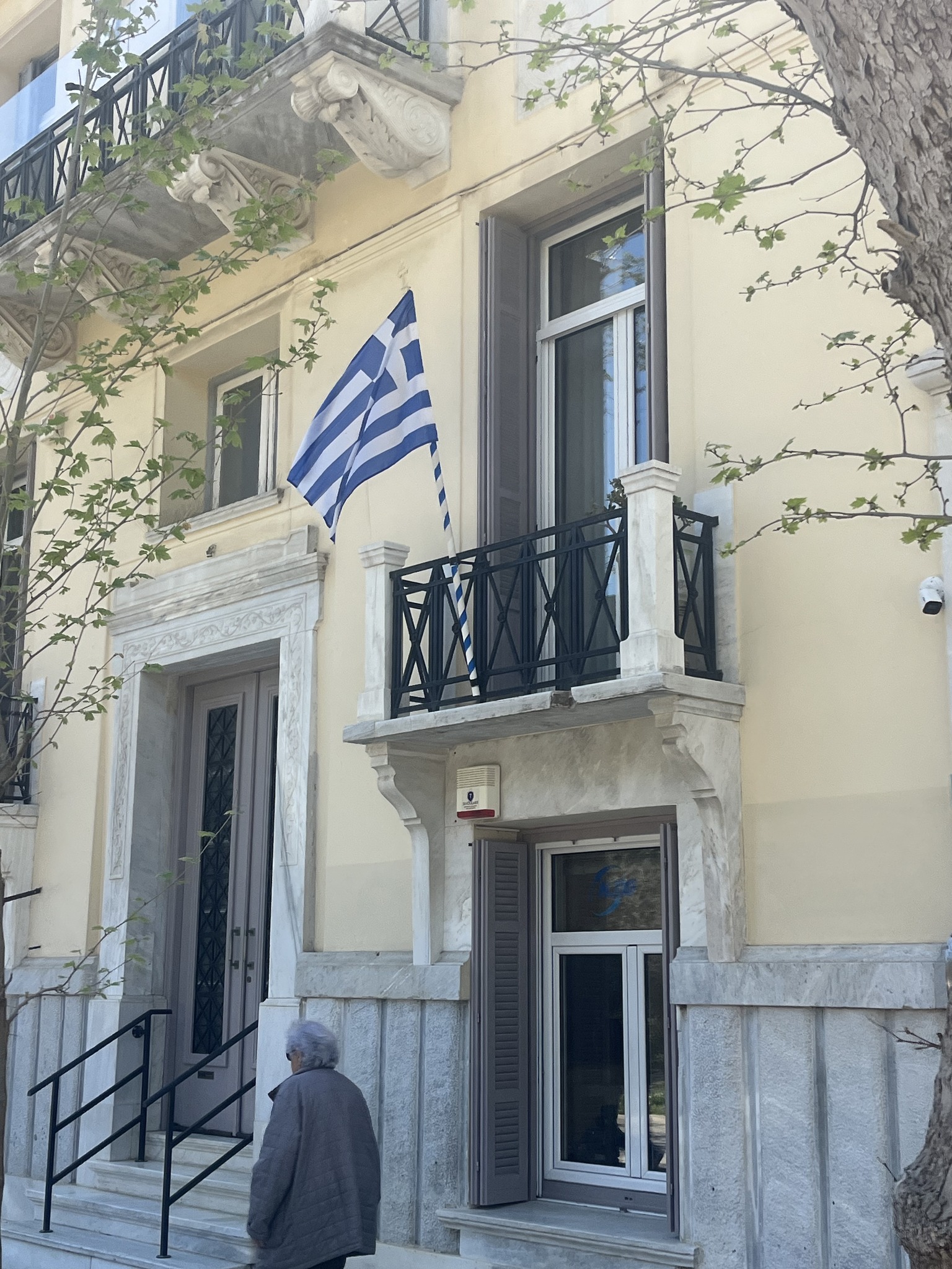 A beige building with a Greek flag on a balcony, two large windows, a marble entrance, and a person standing near the door.