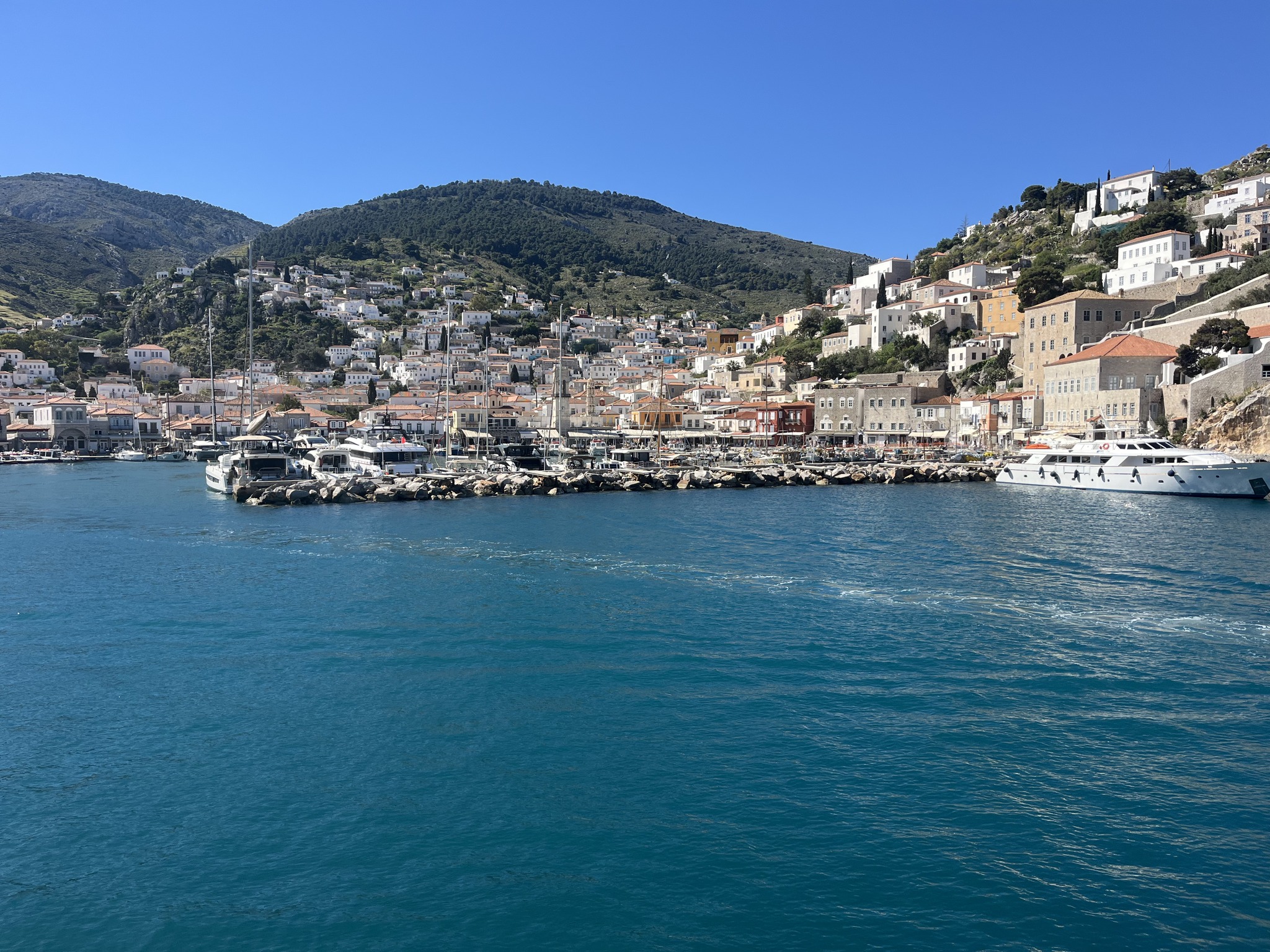 A coastal town with white buildings on hills, boats docked along the waterfront, and clear blue waters under a bright, sunny sky.