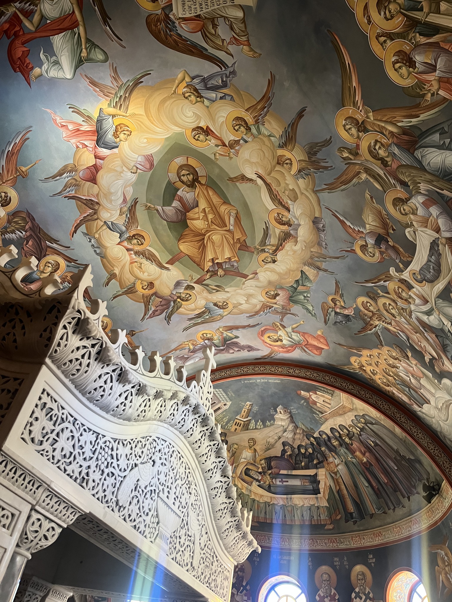 Ornate church ceiling with detailed religious fresco, featuring angels, a central haloed figure, and intricate decorative elements above a white carved staircase.