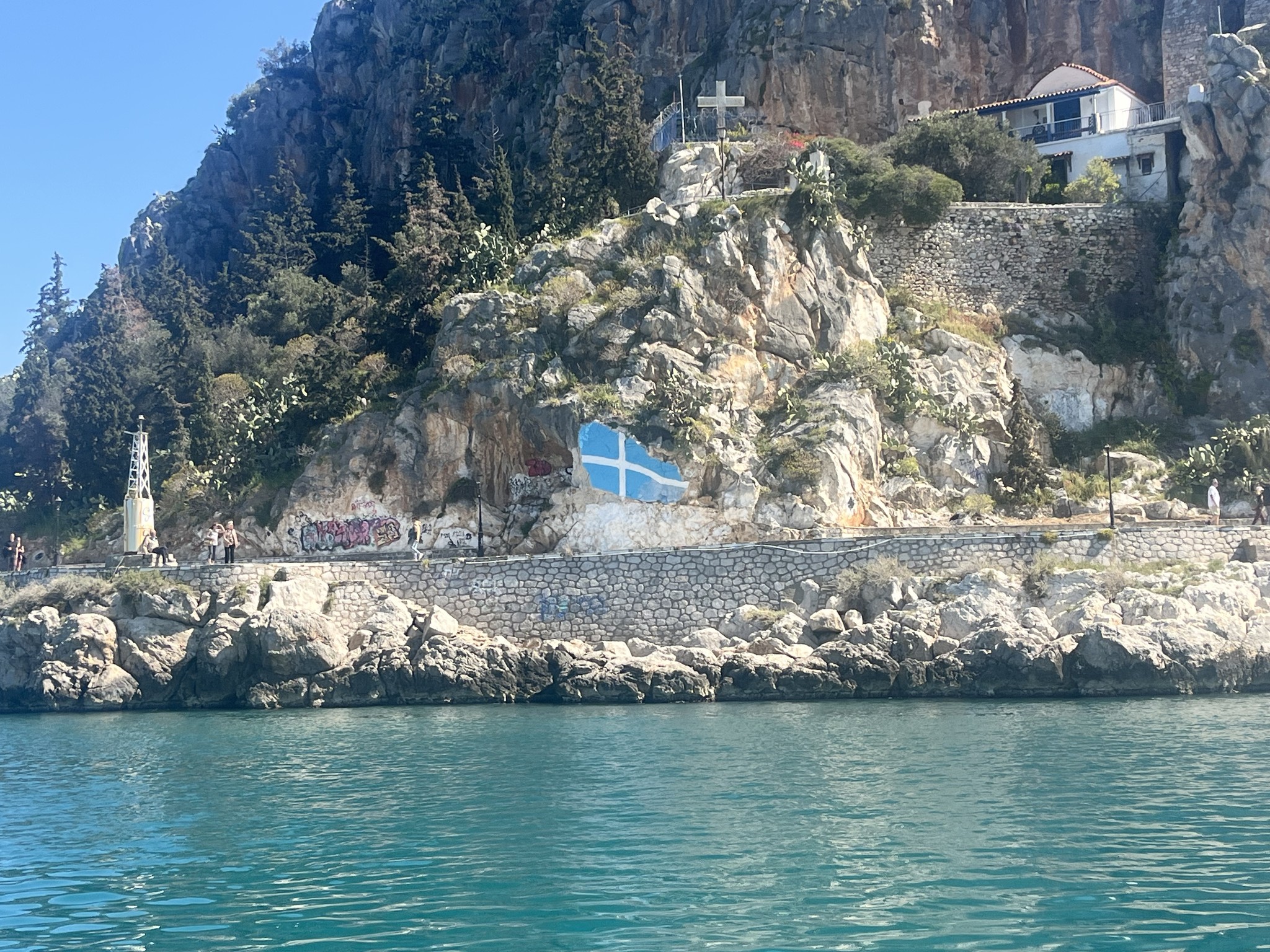 Rocky shoreline with turquoise water, a blue building set into the cliff, greenery, and a small house on top of the hill with a visible cross.