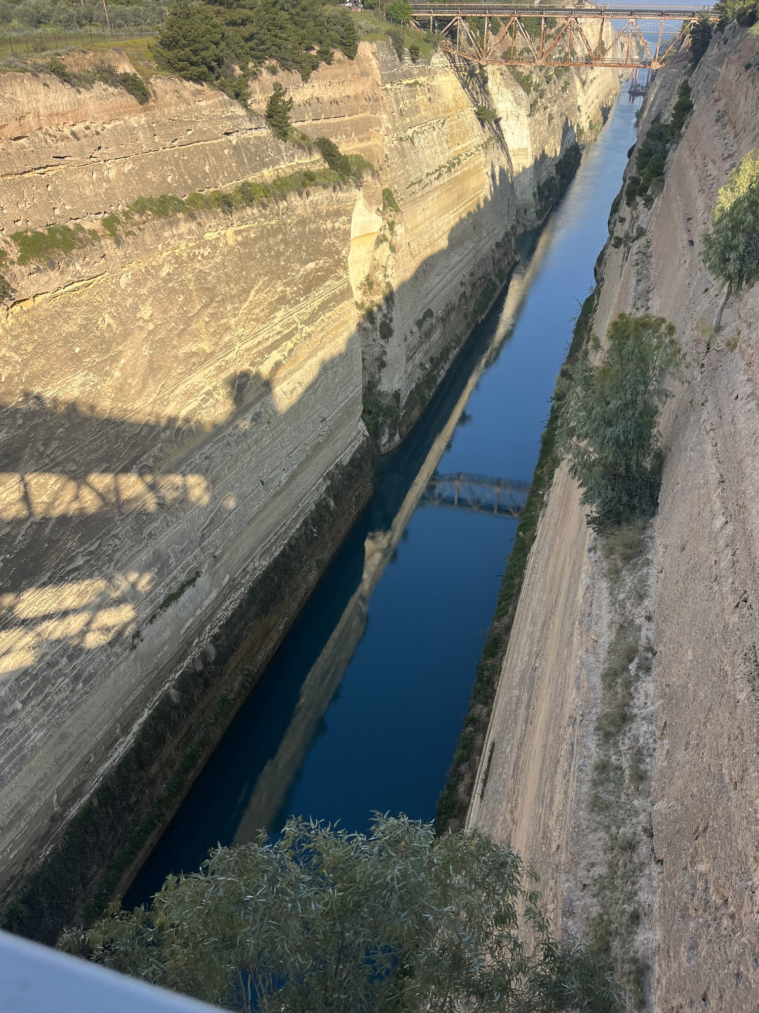 Narrow canal with steep rocky walls, deep blue water, and a bridge casting a shadow overhead. Sparse vegetation lines the edges of the canal.