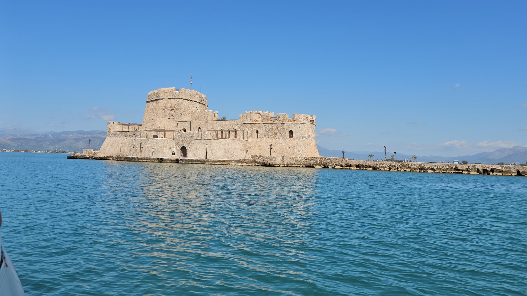Old stone fortress on the water under a clear blue sky, with calm sea in the foreground and a few people walking nearby.