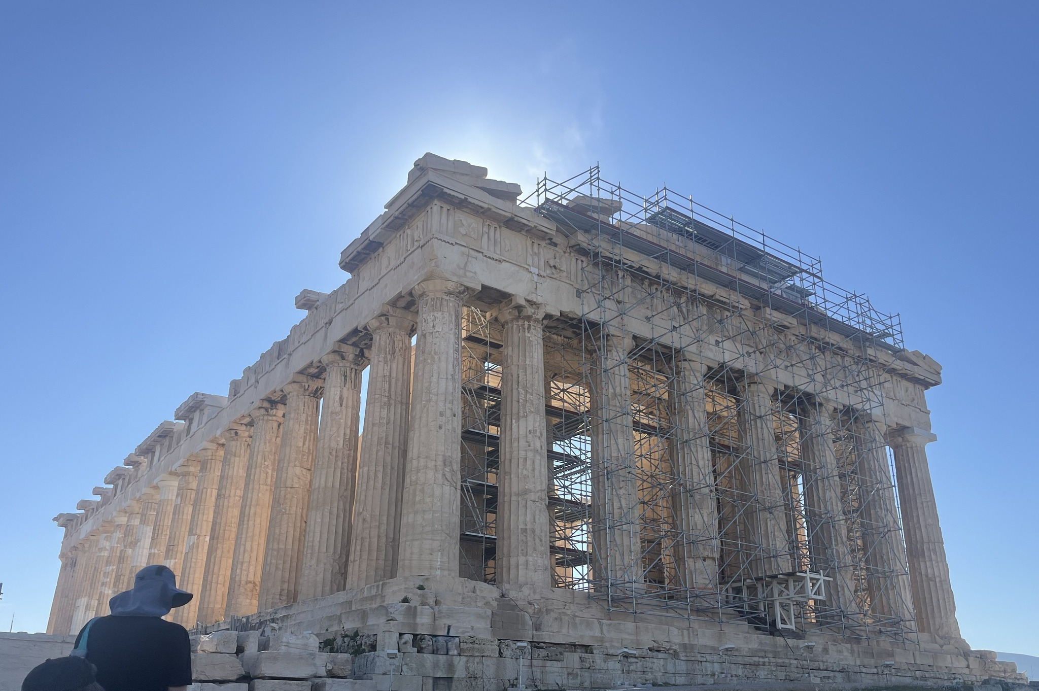 Ancient Greek Parthenon temple with columns and scaffolding under a clear blue sky, sunlight shining behind the structure.