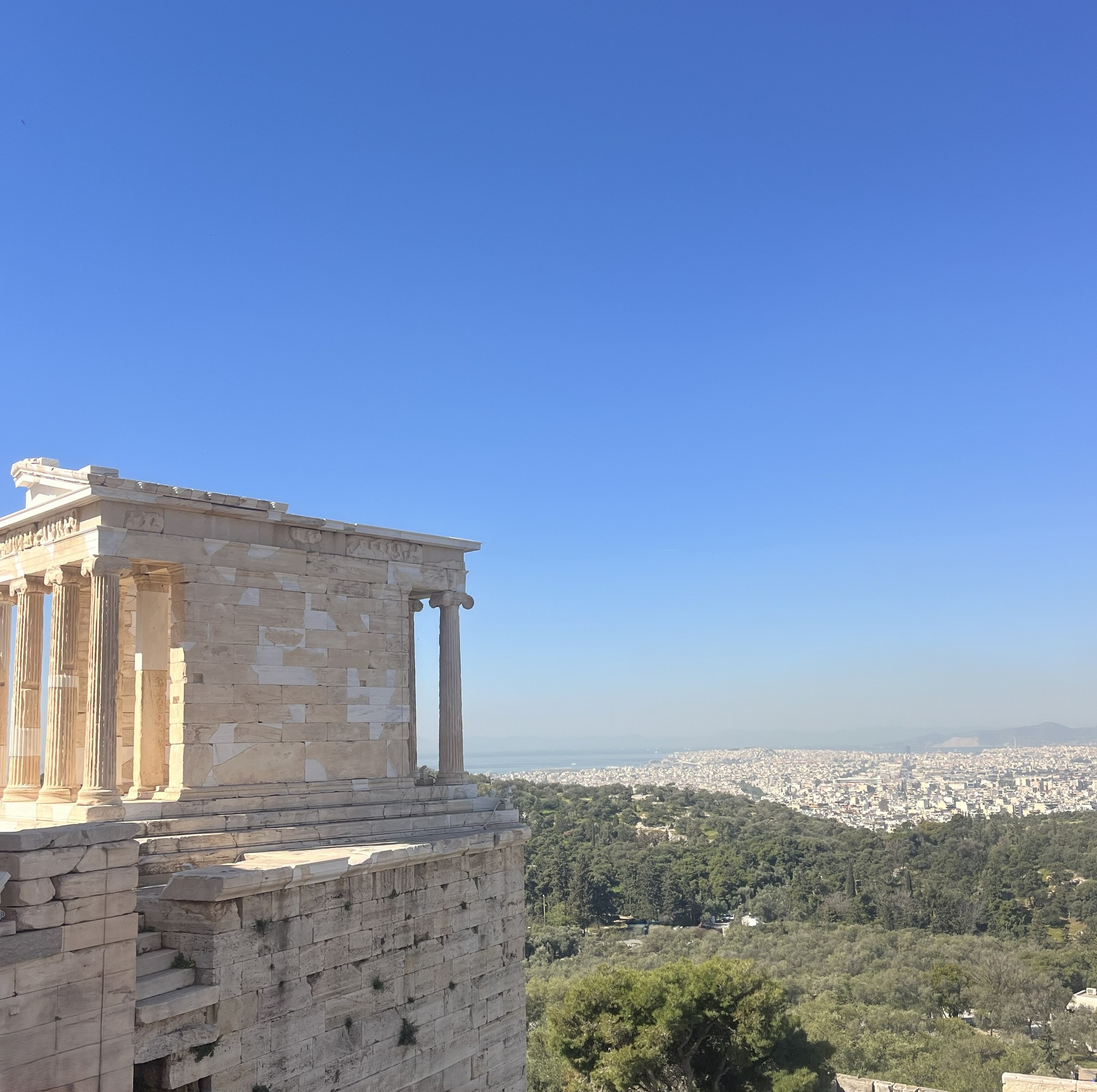 Ancient Greek temple ruins on a hill under a clear blue sky, overlooking a cityscape and green landscape in the distance.