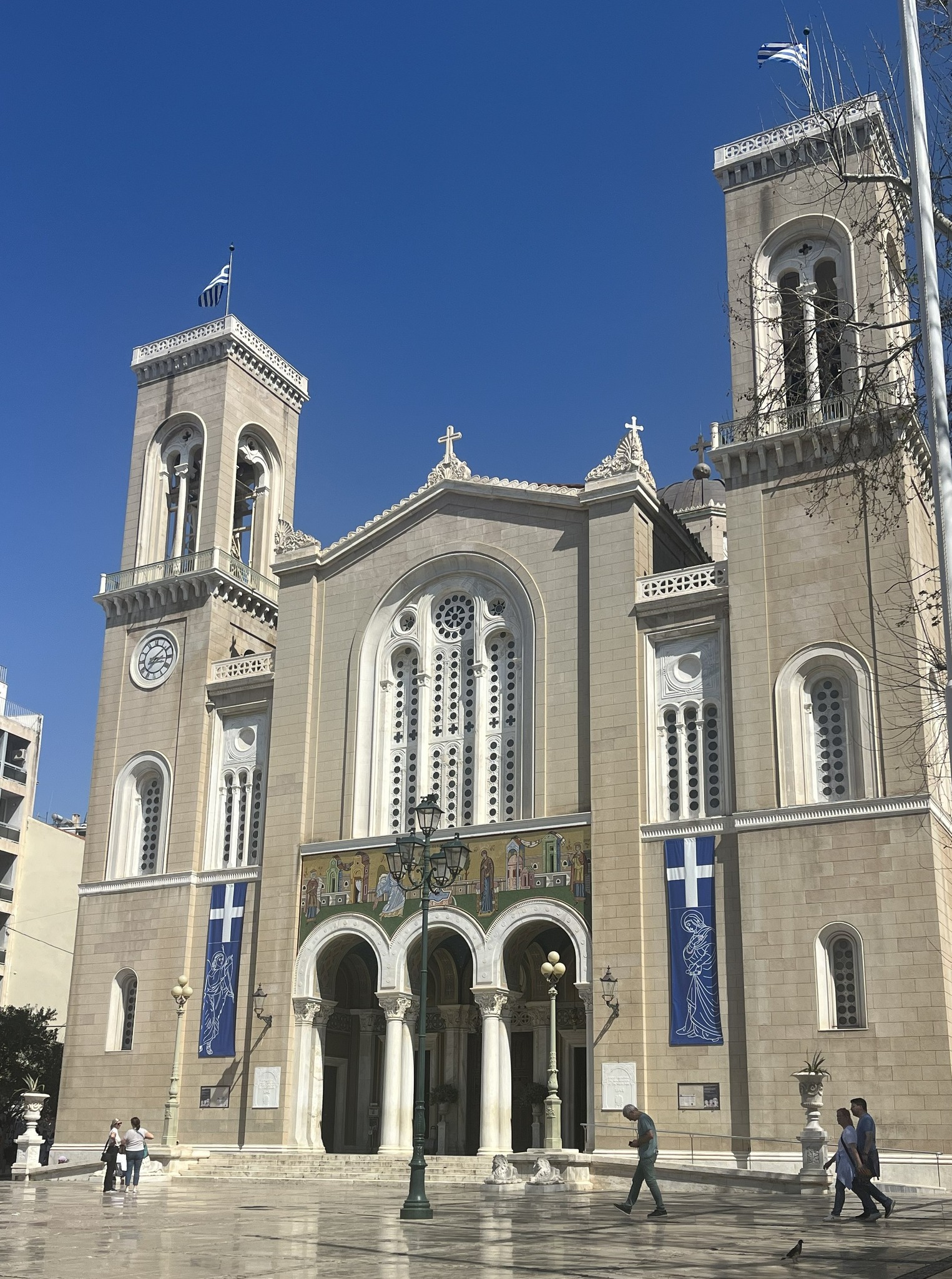 Large stone church with two bell towers, arched windows, and blue banners featuring white crosses, set against a clear blue sky.