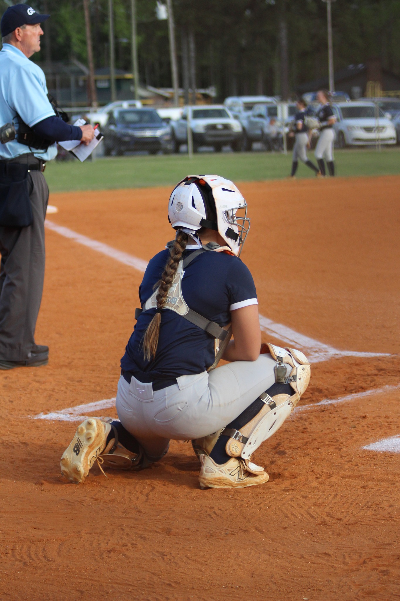 A softball catcher in full gear crouches behind home plate, ready to receive a pitch during a game, with an umpire standing nearby.
