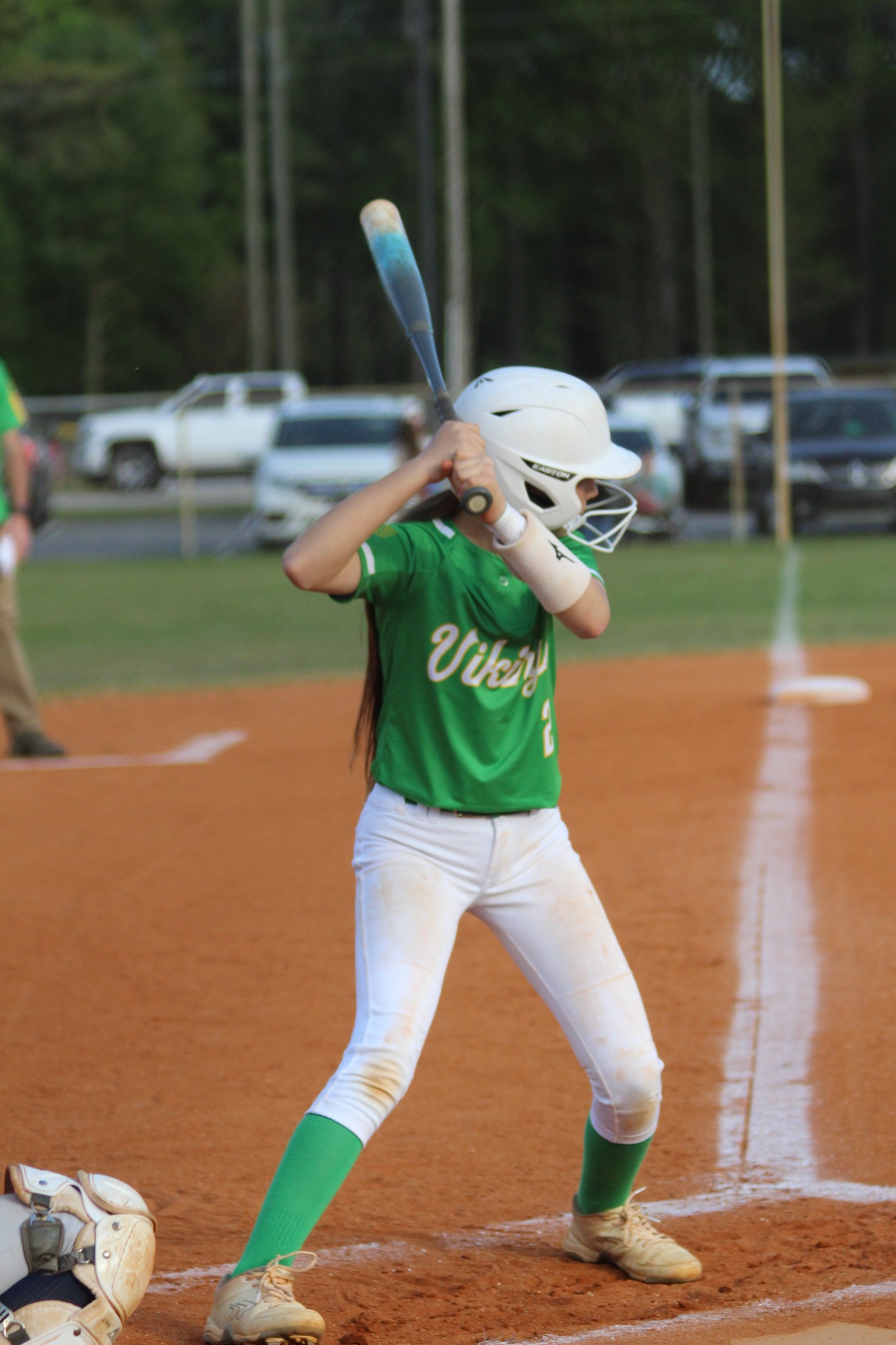 A young softball player in a green shirt and white helmet prepares to bat on a dirt field, with cars and people in the background.