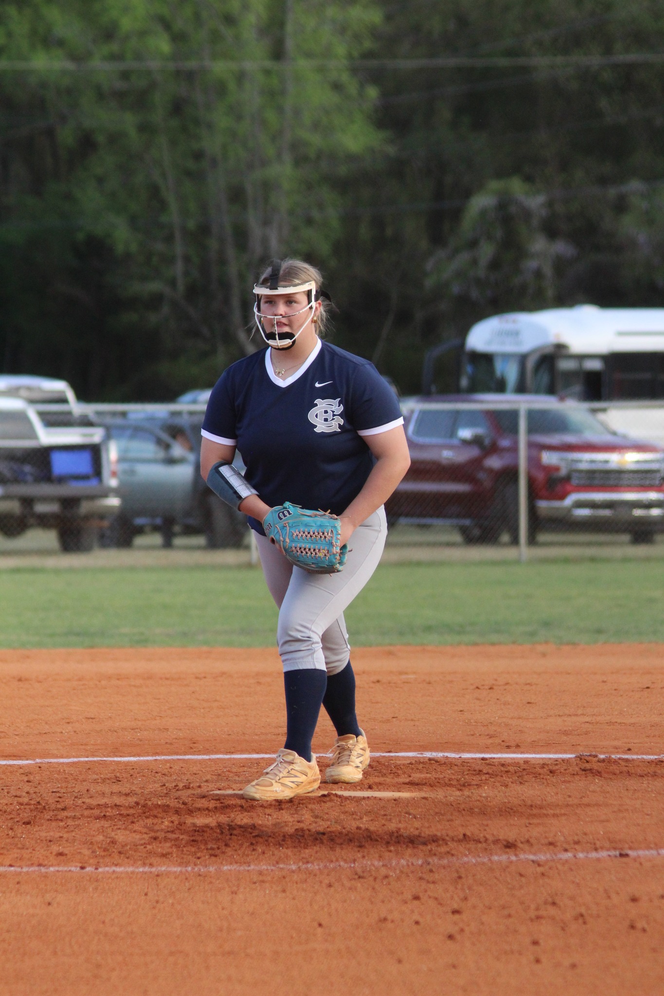 A softball player in uniform stands on the pitcher's mound, preparing to pitch during a game. Trees and parked cars are visible in the background.